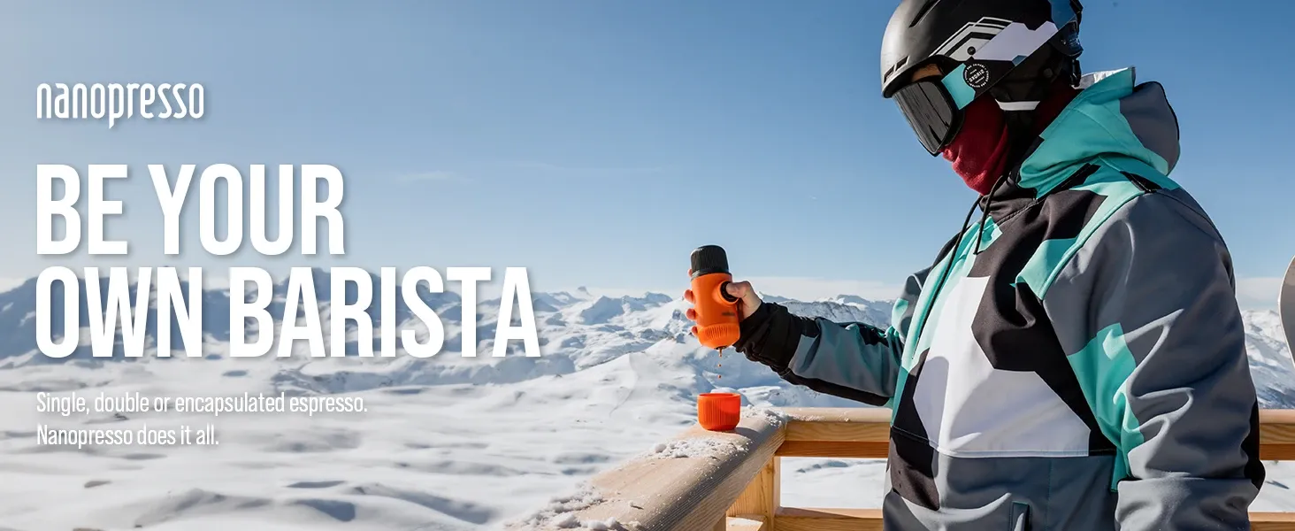 A person in ski gear using a bright orange Nanopresso manual espresso maker on a snowy mountain balcony with a panoramic Alpine background.